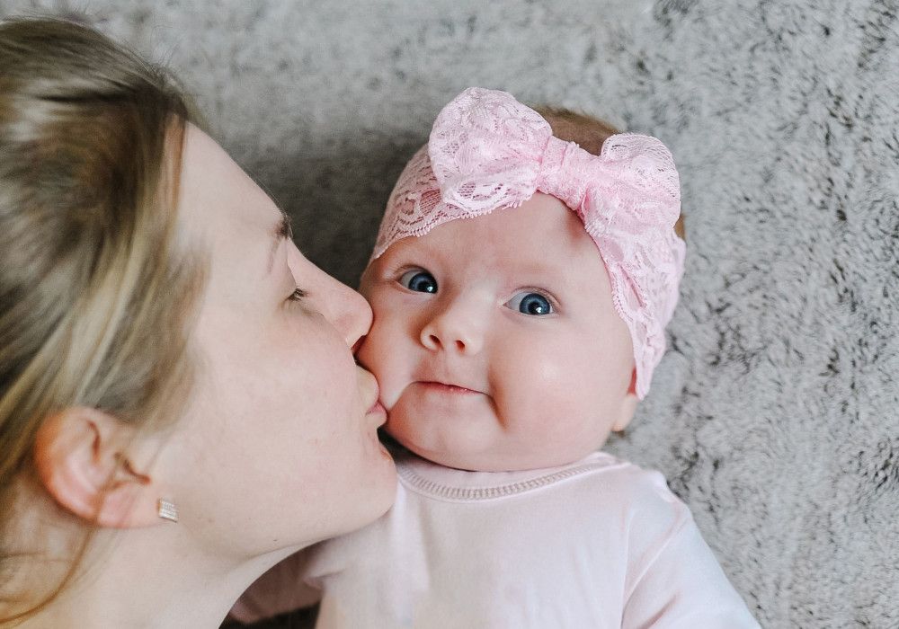 Una madre dando un beso a su bebé. La niña tiene un lazo grande rosa en la cabeza. Cuidado infantil.
